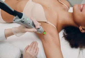 Close-up of technician applying green-tipped laser device to client's underarm while assisting hand steadies area in a professional medical aesthetics laser training procedure.