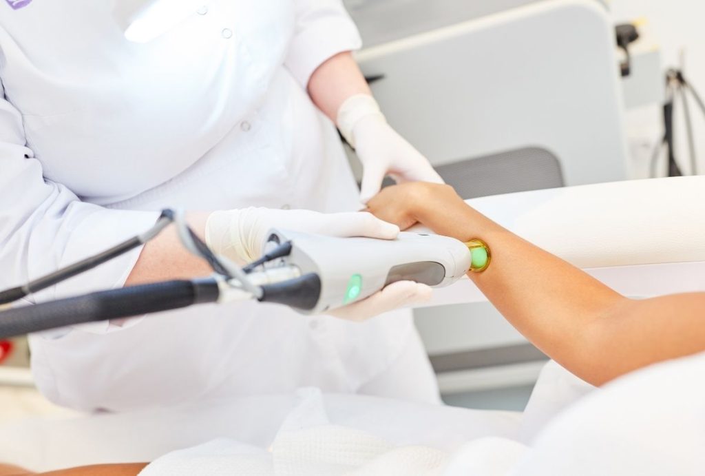 Gloved technician applying green-lit laser device to client's arm while stabilizing with hand during esthetician laser training program session in modern clinic.
