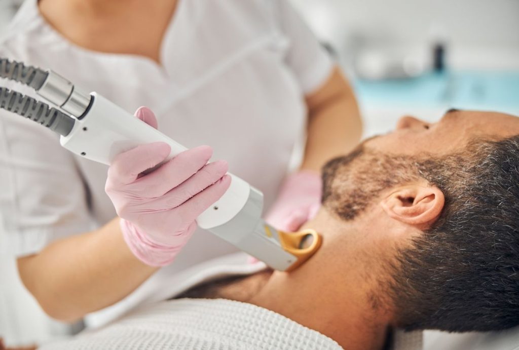 Technician with pink gloves performing neck laser hair removal on male client lying relaxed in a clinic for medical aesthetics laser training session.